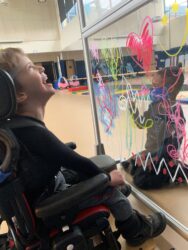 Young boy in wheelchair laughs at another boy on other side of decorated plexi-glass barrier