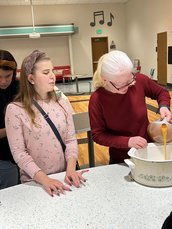 On student reads the recipe for spiced apple cider in Braille while another student adds ingredients to the slow cooker.