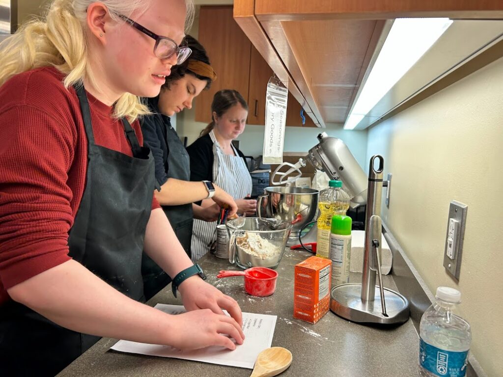 A student reads the recipe for pumpkin bars in Braille while another student measures ingredients and a teacher instructs in non-visual baking methods.