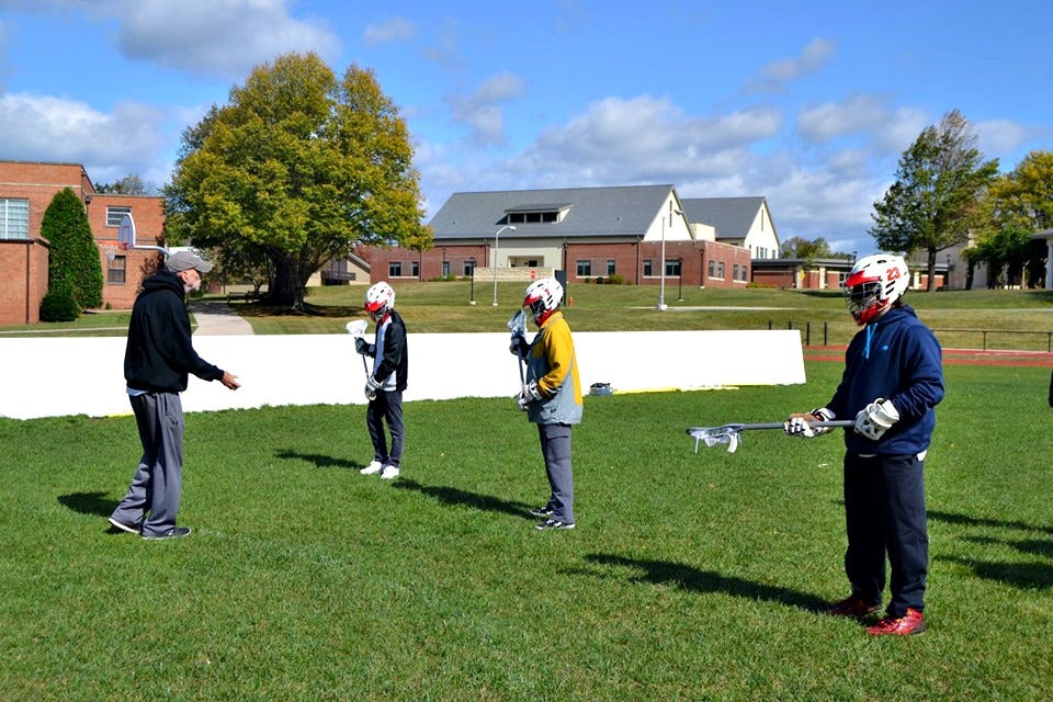Students stand with coaches learning how to hold a lacrosse stick.