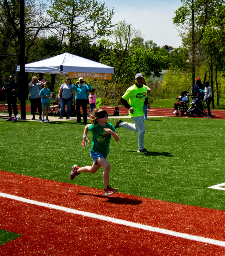 Kid runs towards beep baseball bas wearing eyeshades, as a volunteer runs along and cheers him on.