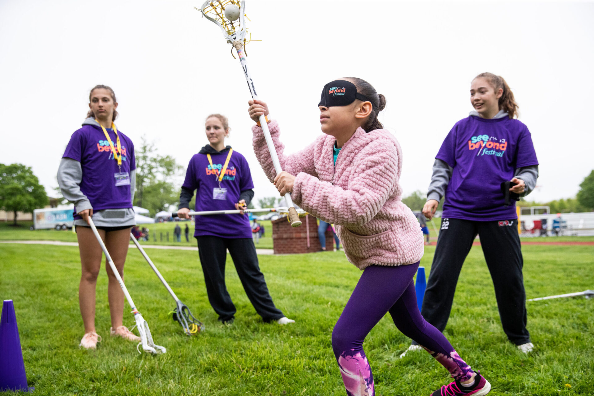a group of young girls instructing a girl wearing eyeshades on throwing a lacrosse ball from the stick . a group of young girls instructing a girl wearing eyeshades on throwing a lacrosse ball from the stick .