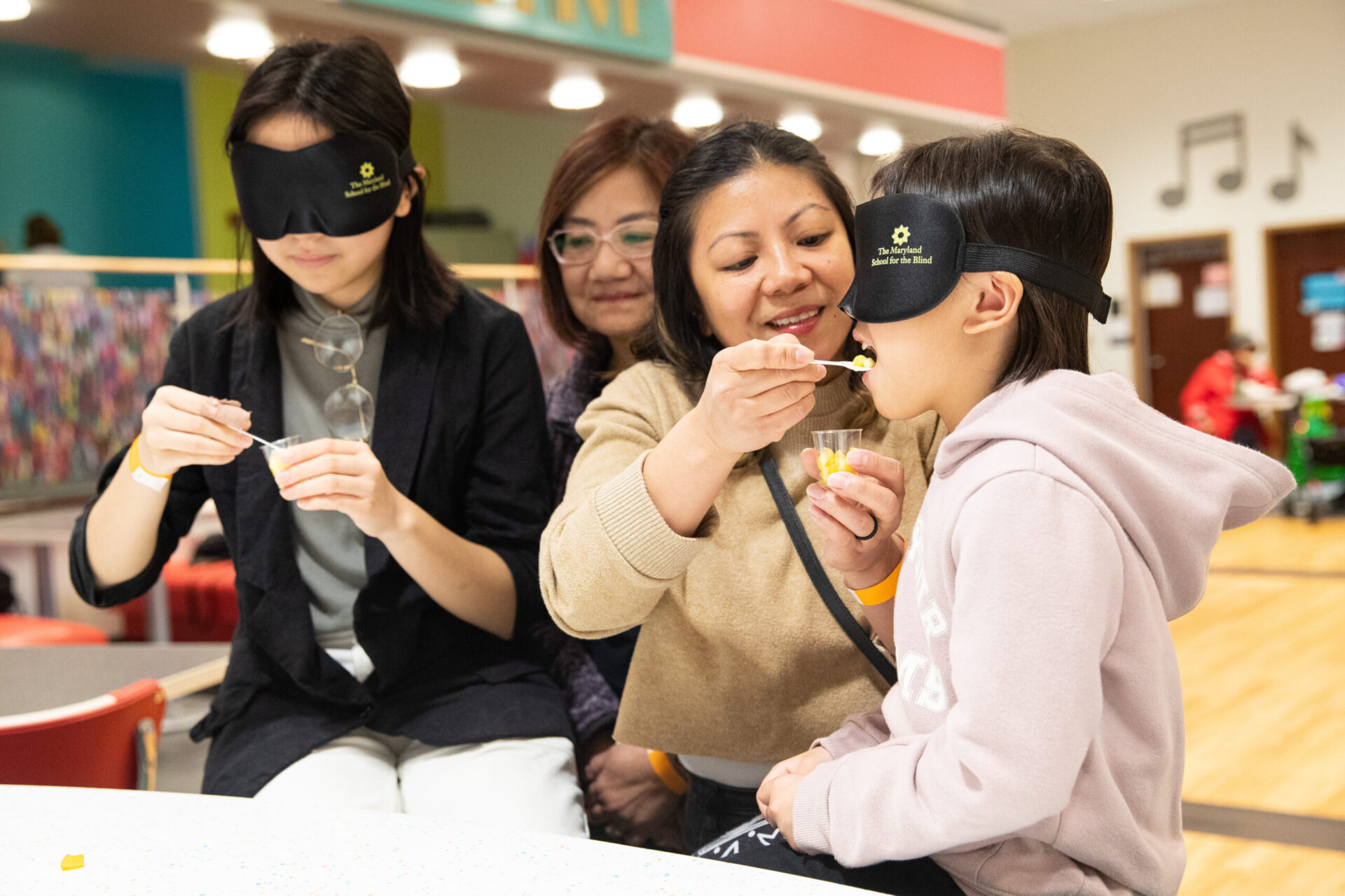 Two individuals with blindfolds sit as a woman helps a child taste food using a spoon. Another person observes in the background. The setting is an indoor space with colorful decor. Two individuals with blindfolds sit as a woman helps a child taste food using a spoon. Another person observes in the background. The setting is an indoor space with colorful decor.
