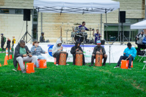 a group of students seated in front of a stage, drum on buckets and Djembe drums.