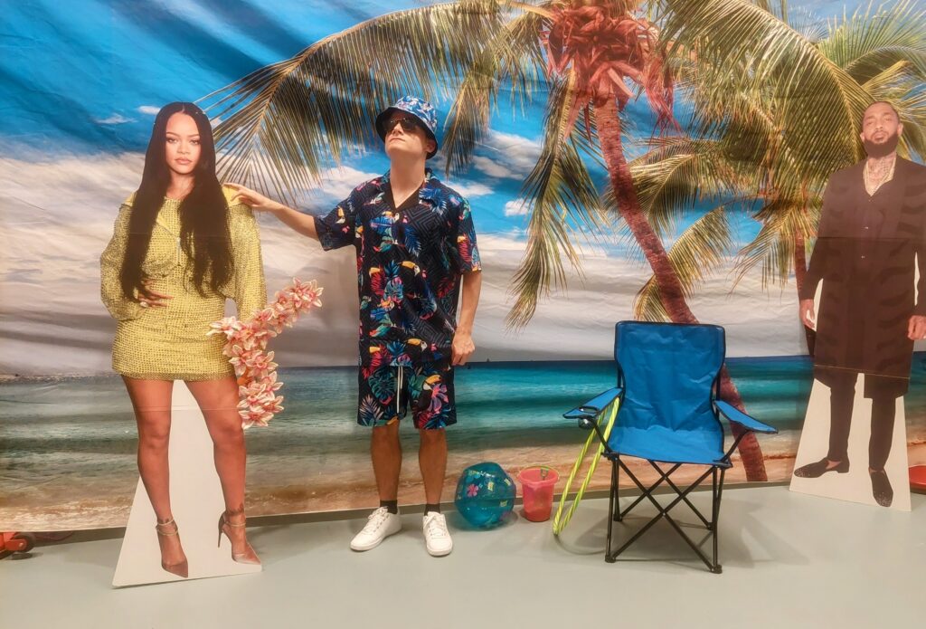 A man and a cardboard woman standing next to a chair with a palm tree in the background.