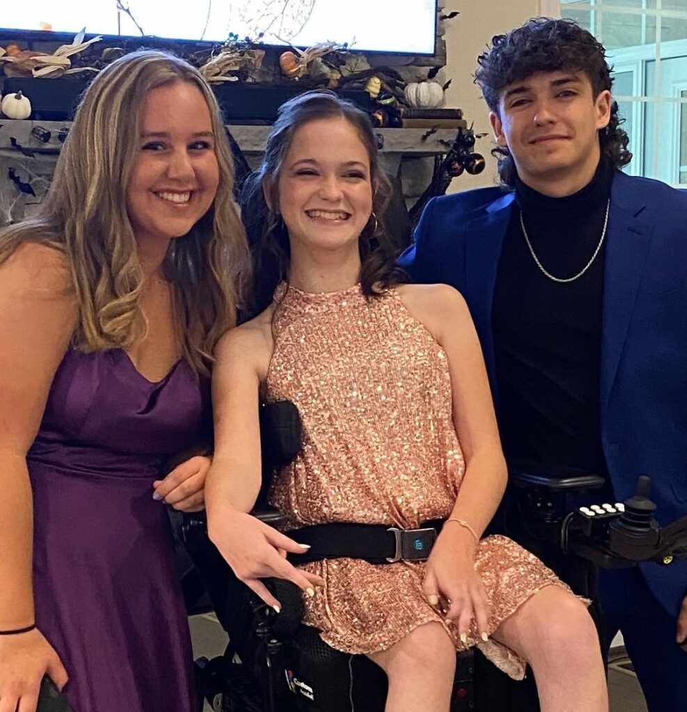 Three young people dressed for a formal occasion, with one person in a wheelchair in the center. All are smiling and posing for the photo.