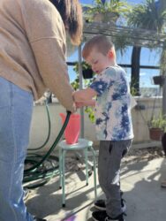 Miss Jenn shows a student, Connor, how to use a garden hose to fill a watering can.