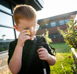 A young student looks at a flower close to his classes. He is outdoors in the Early Learning Greenhouse Classroom.