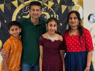 a family of four standing together, smiling for the camera. In the center, the father, wearing a green polo shirt and jeans, has his arms around his daughter and son. The daughter is dressed in a maroon lace dress and accessorized with jewelry, while the son wears an orange and white striped shirt with black pants. The mother, on the far right, is dressed in a red and navy blue traditional outfit with gold detailing.