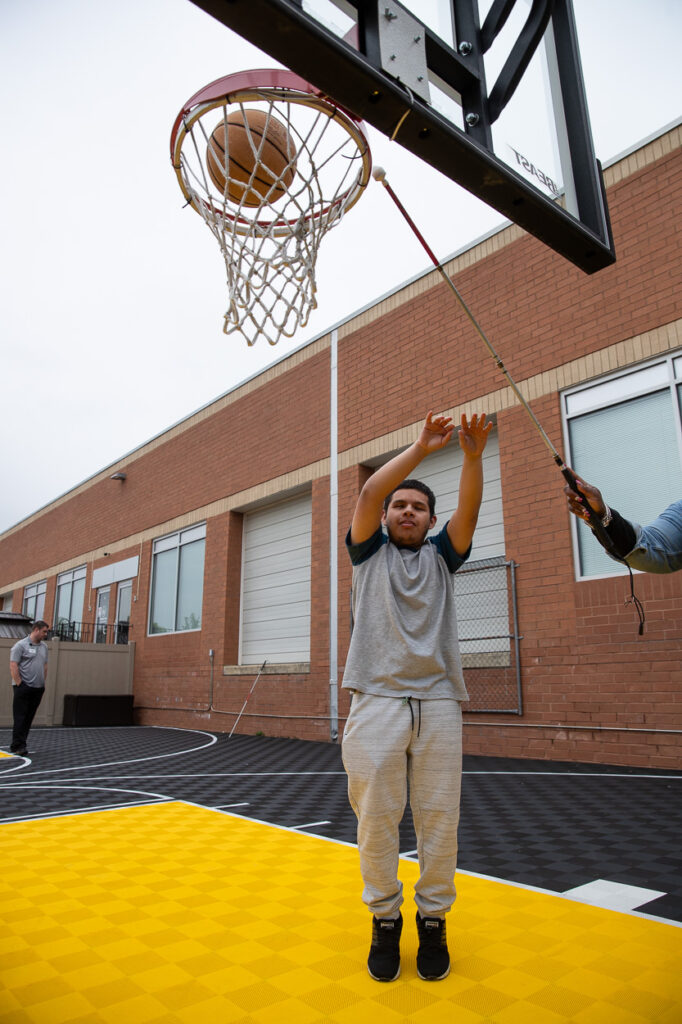 A young person in gray athletic wear shoots a basketball toward a hoop while someone else uses a pole to hold the net steady on an outdoor court with a brick building in the background.