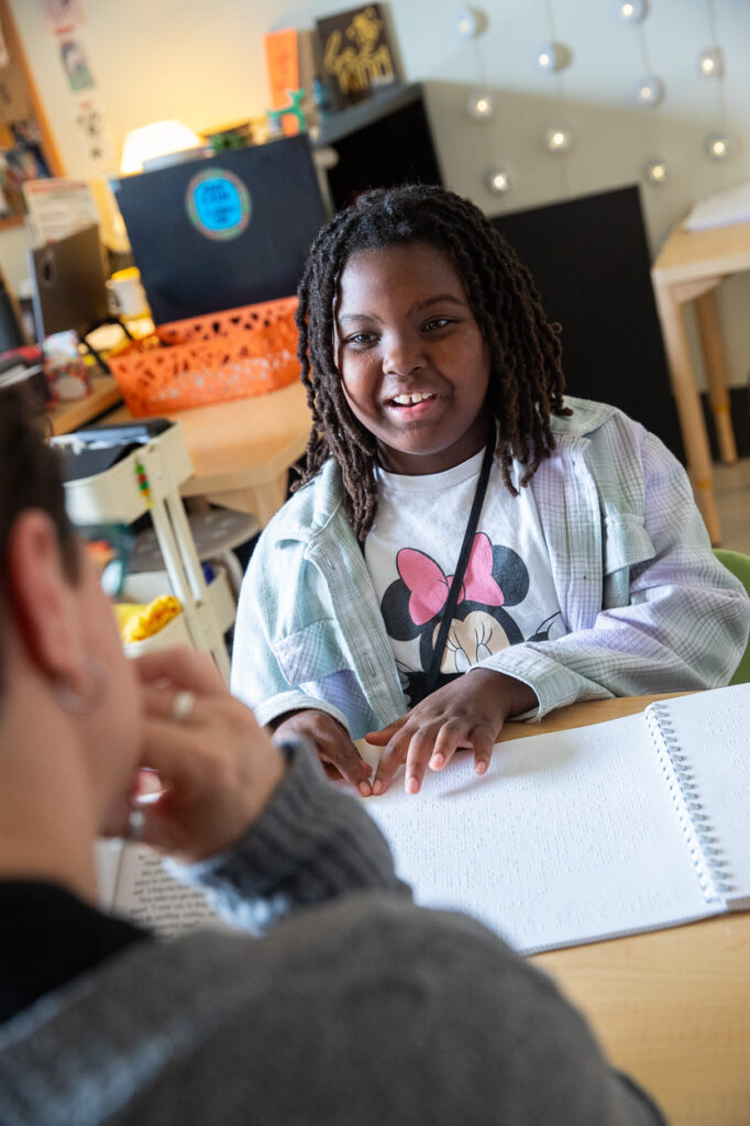 A young girl with shoulder-length locs, wearing a light plaid shirt and a Minnie Mouse t-shirt, smiles while sitting at a table, reading Braille from a notebook as she interacts with another person.