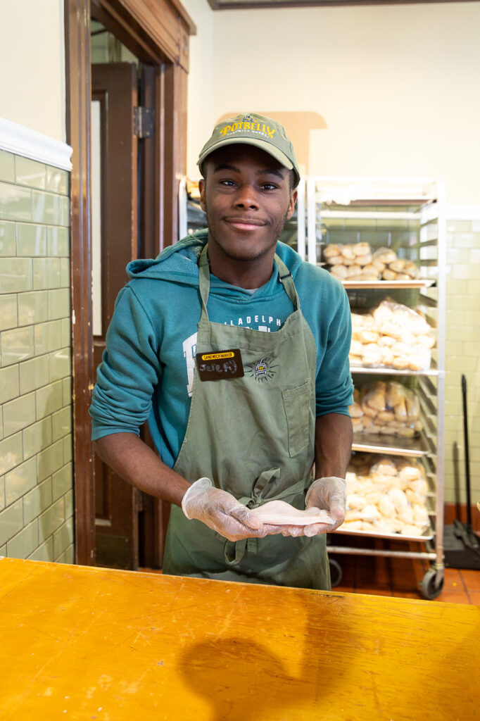 A young man wearing a green apron, cap, and gloves stands behind a counter, smiling and holding a piece of dough. Behind him are shelves filled with bread rolls in a bakery setting.