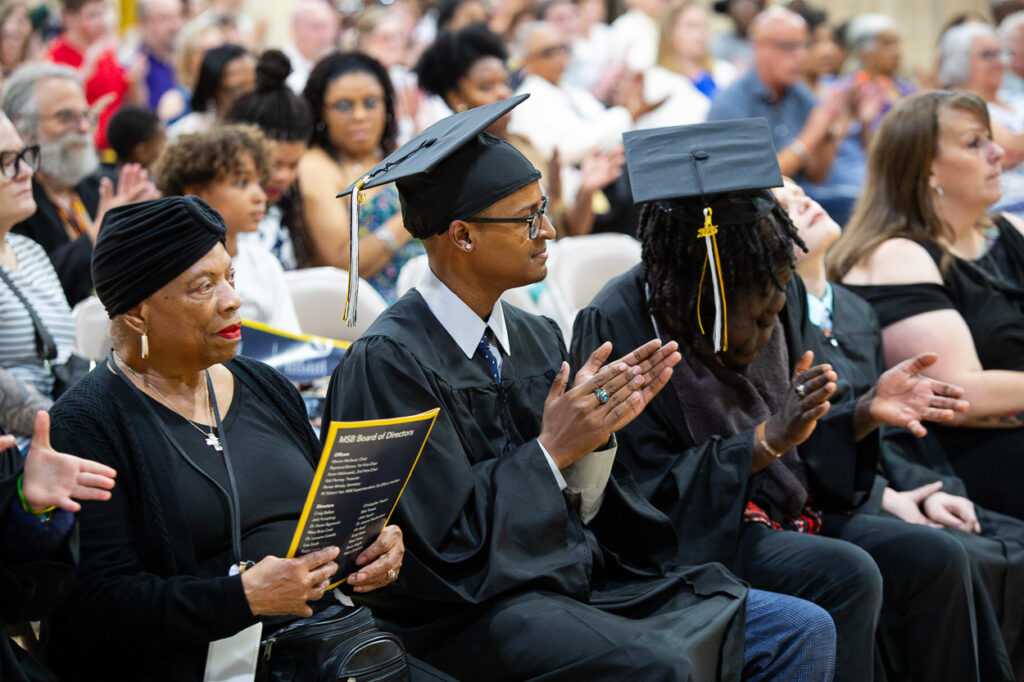 At our 2025 graduation ceremony, a group of attendees is gathered, sharing in the joy of this special day. Among them, two proud graduates in their caps and gowns applaud as they celebrate this milestone. Beside them, an older woman holds the program in her hands, all smilesâtogether capturing a memorable scene that perfectly embodies the spirit of achievement and community for our Summer Newsletter.