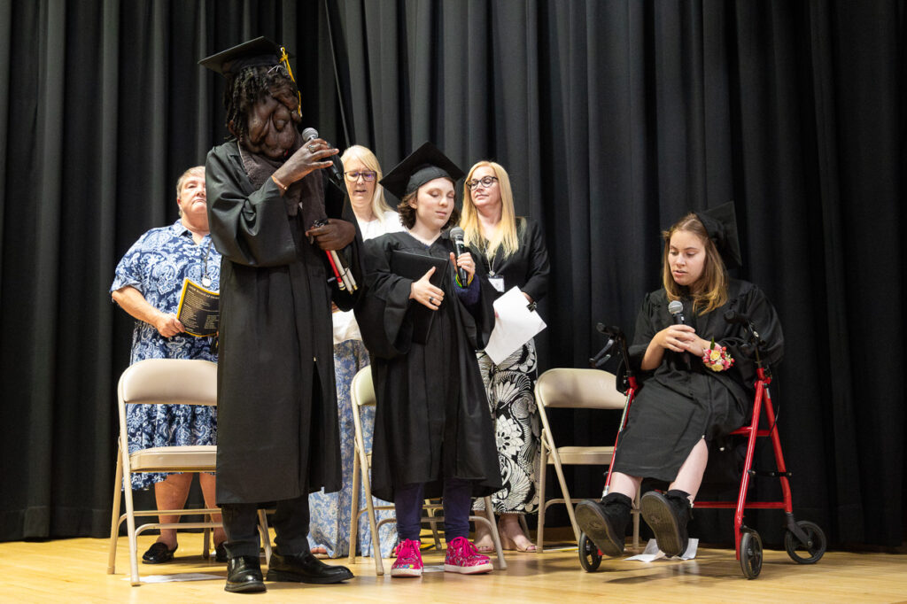 At our 2025 Summer Graduation Ceremony, three proud graduatesâdressed in caps and gowns, including one using a walkerâstand confidently on stage with microphones in hand. Behind them, four supportive adults look on, all set against a black curtain backdrop. This special moment is featured in the latest edition of The Maryland School for the Blind's 2025 Summer Newsletter.