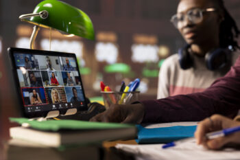 A person joins a video conference on a tablet at a desk cluttered with books, papers, and pens, drawing inspiration from the lively atmosphere. Nearby, someone with headphones around their neck sits under a green desk lamp.