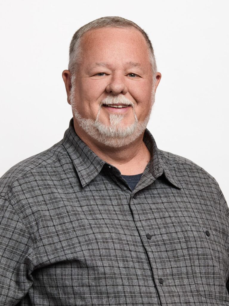 A middle-aged man with short gray hair and a gray beard, wearing a gray plaid button-up shirt, smiles in front of a plain white background, embodying the approachable professionalism of MSB Leadership.