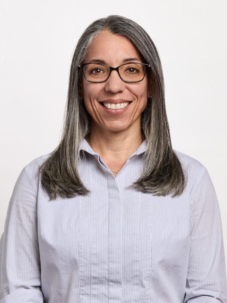 A woman with long, straight gray hair and glasses smiles at the camera, embodying MSB Leadership. She is wearing a light blue, button-up shirt and is posed against a plain white background.