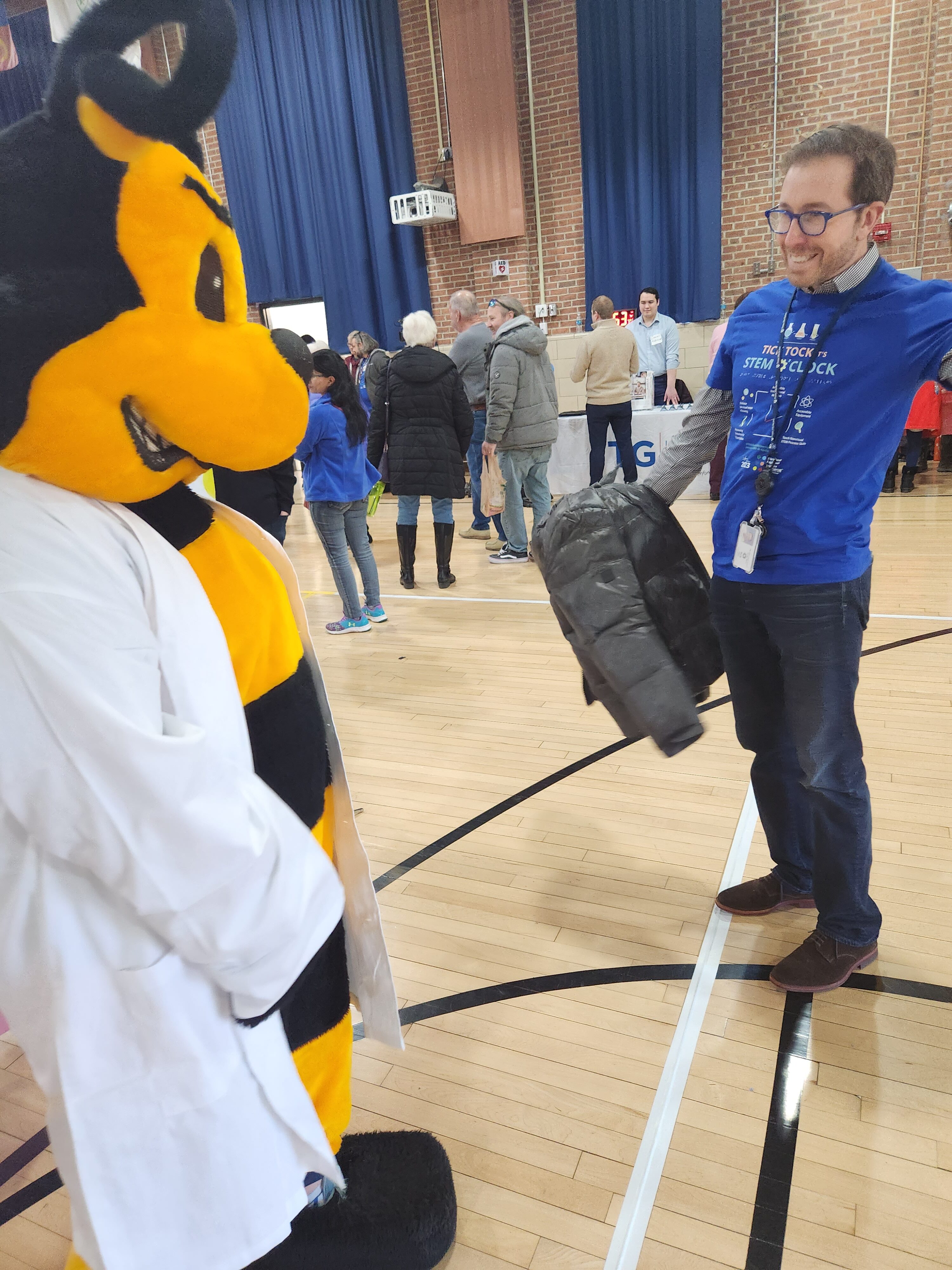 A person in a blue shirt and glasses holds out a jacket while interacting with someone in a bee mascot costume wearing a white lab coat at an indoor event with other people in the background. A person in a blue shirt and glasses holds out a jacket while interacting with someone in a bee mascot costume wearing a white lab coat at an indoor event with other people in the background.