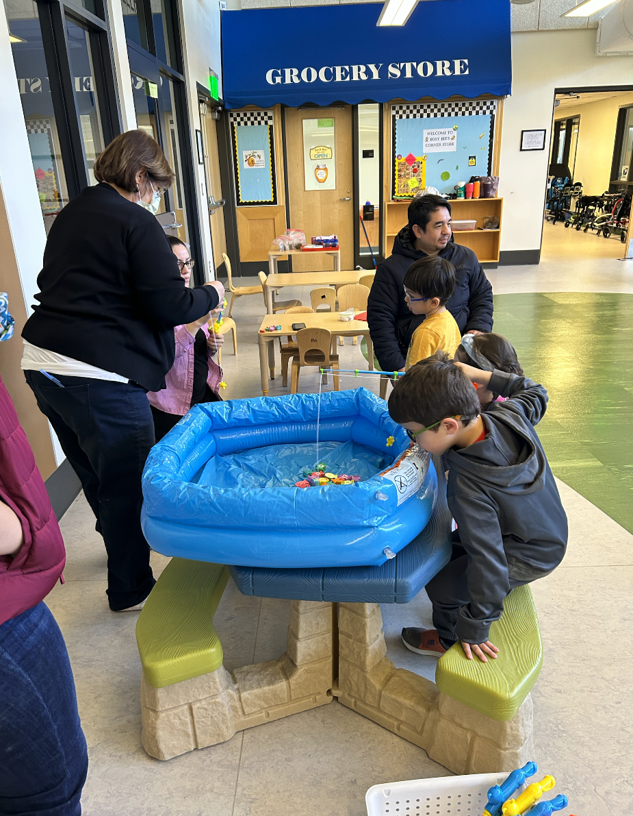 Children and adults gather around a small blue inflatable pool filled with toy fish in a play area. The setting resembles a classroom or daycare with a pretend grocery store in the background. Children and adults gather around a small blue inflatable pool filled with toy fish in a play area. The setting resembles a classroom or daycare with a pretend grocery store in the background.