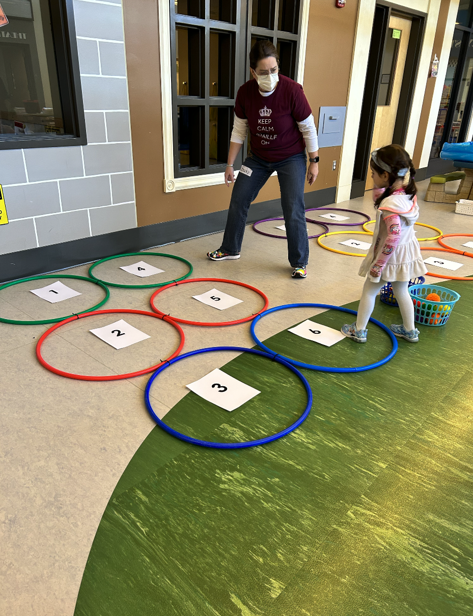 A woman and a young girl play an educational hopping game using numbered papers inside colorful hoops arranged on the floor indoors. The woman appears to guide the girl as she hops from one hoop to another. A woman and a young girl play an educational hopping game using numbered papers inside colorful hoops arranged on the floor indoors. The woman appears to guide the girl as she hops from one hoop to another.