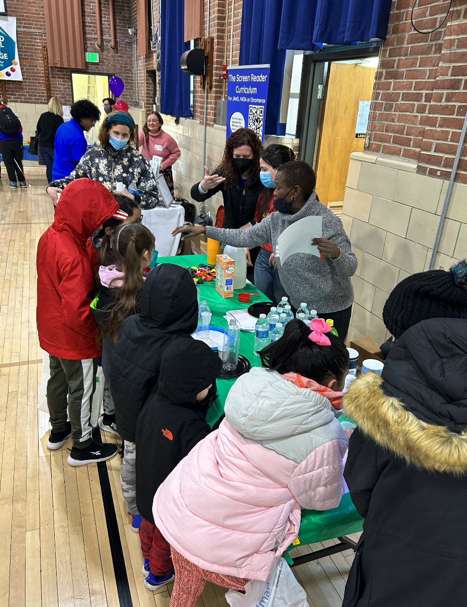 A group of children and adults gather around a table with drinks and colorful items at an indoor event, likely in a school gym, while a woman speaks and others observe. Some people wear masks and winter jackets. A group of children and adults gather around a table with drinks and colorful items at an indoor event, likely in a school gym, while a woman speaks and others observe. Some people wear masks and winter jackets.