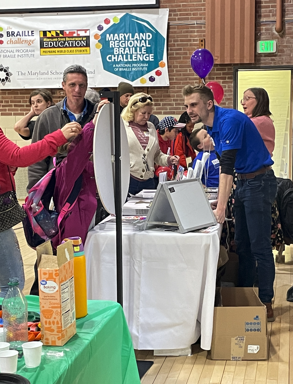 People gather around a table at an event, with informational materials and laptops on display. A “Maryland Regional Braille Challenge” banner hangs above. Balloons and drinks are visible, creating a lively and welcoming atmosphere. People gather around a table at an event, with informational materials and laptops on display. A “Maryland Regional Braille Challenge” banner hangs above. Balloons and drinks are visible, creating a lively and welcoming atmosphere.