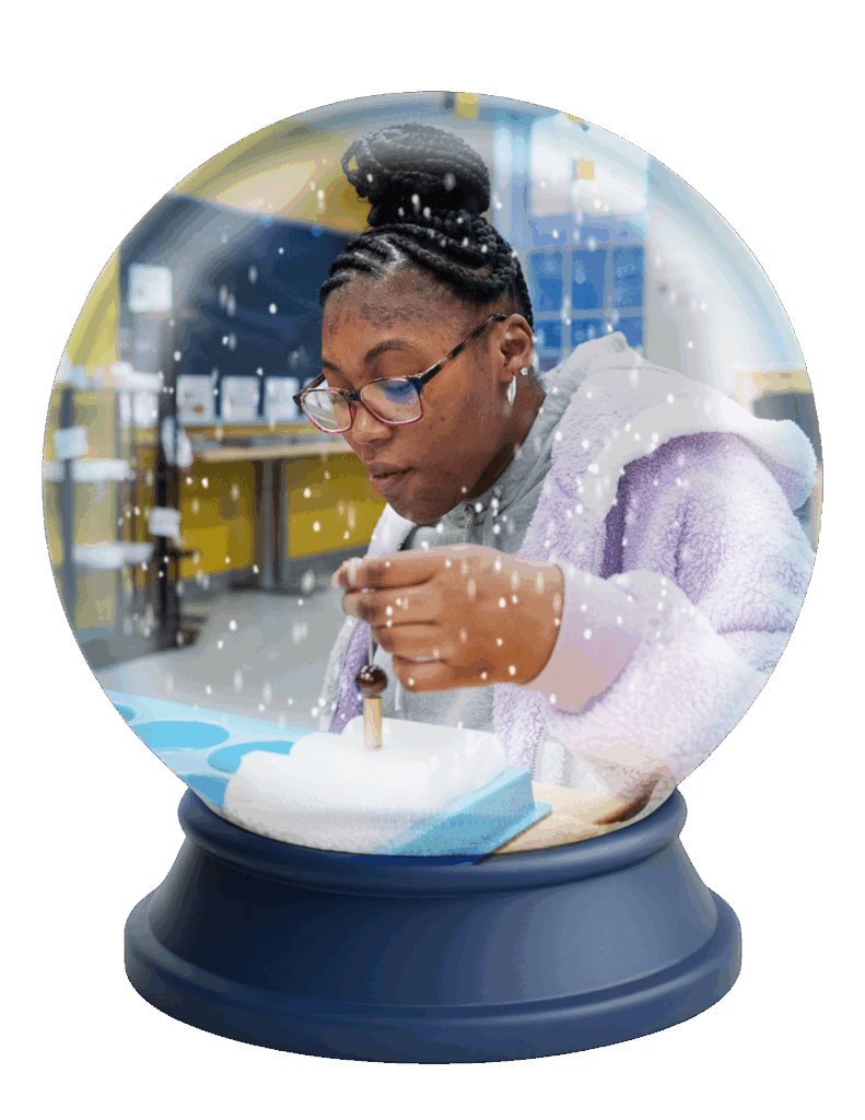 A young woman in glasses focuses on a project inside a classroom, as if she is inside a snow globe with falling snow and a Kindle-blue base, embodying the Gift of Communication.