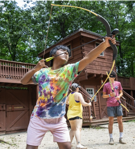 A teenager in a tie-dye shirt aims a bow and arrow outdoors, with two other people and a wooden cabin in the background. Trees and greenery surround the scene, suggesting a summer camp setting.