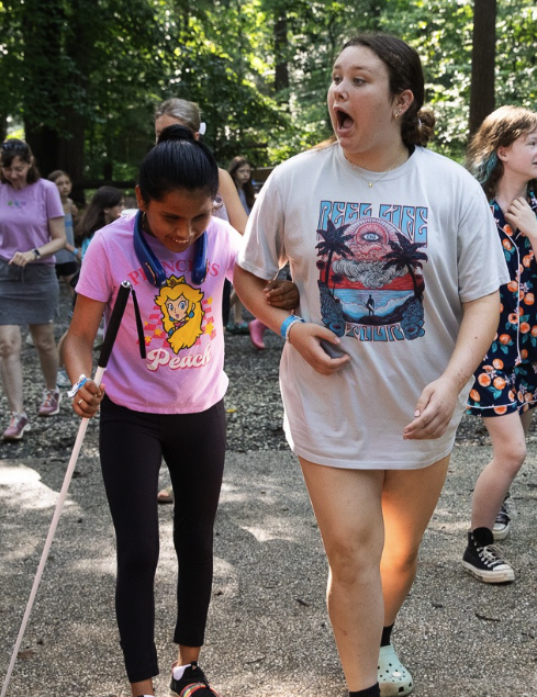A girl wearing a Princess Peach shirt and holding a white cane walks arm-in-arm with another girl in a large T-shirt, both smiling and laughing, surrounded by other people outdoors in a wooded area.