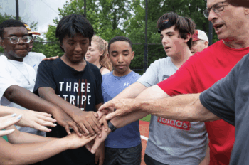 A diverse group of children and an adult stand in a circle outdoors, putting their hands together in the center in a gesture of teamwork and unity, on a sports field with trees in the background.