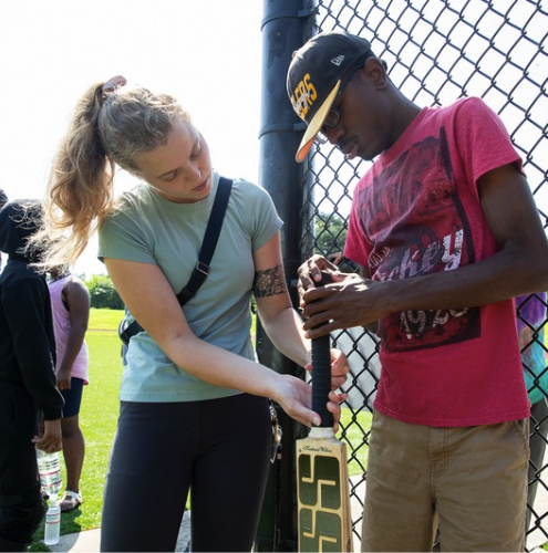 A woman helps a man adjust a cricket bat near a chain-link fence on a sunny day, with a grassy field in the background. Other people are visible nearby.