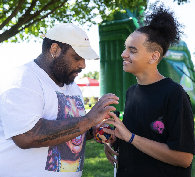 Two people stand outside under trees, smiling at each other. One person, wearing a white cap and shirt, gently touches the other's hands as they hold a colorful ball. The second person has curly hair and wears a black t-shirt.