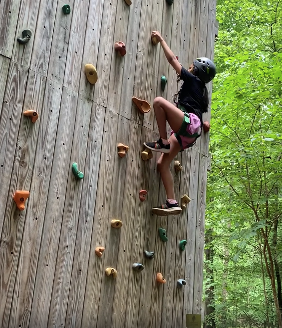 A person wearing a helmet and harness climbs an outdoor wooden rock climbing wall with colorful handholds, surrounded by green trees.