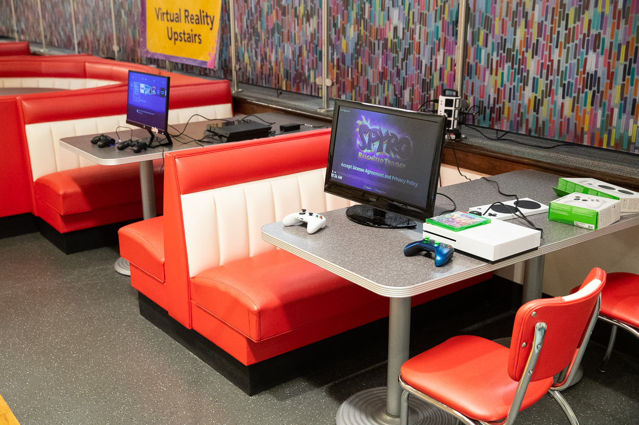 Two red and white diner booths hold video game consoles, controllers, and monitors displaying games. A colorful patterned wall and a yellow “Virtual Reality Upstairs” sign set the scene at the See Beyond Festival.