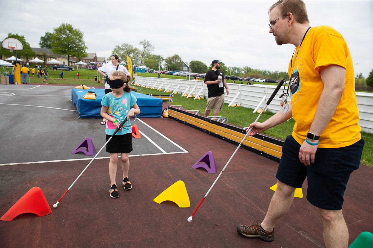 At the See Beyond Festival, a blindfolded child and an adult, both using white canes, navigate a colorful obstacle course on a basketball court, with the adult guiding the child. Other participants and a playground appear in the background.