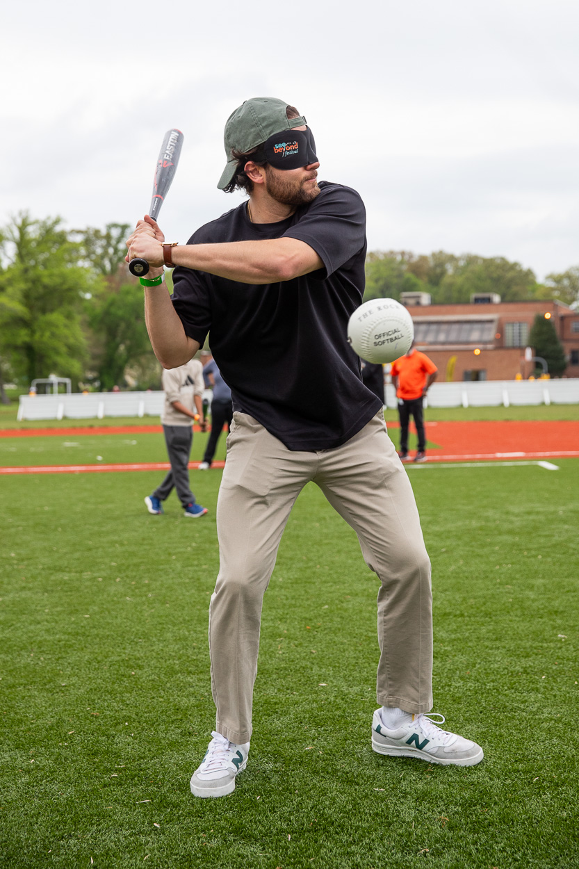 A man wearing a blindfold and a baseball cap prepares to swing a bat at a ball on a grassy field during the See Beyond Festival, with other people and buildings visible in the background.