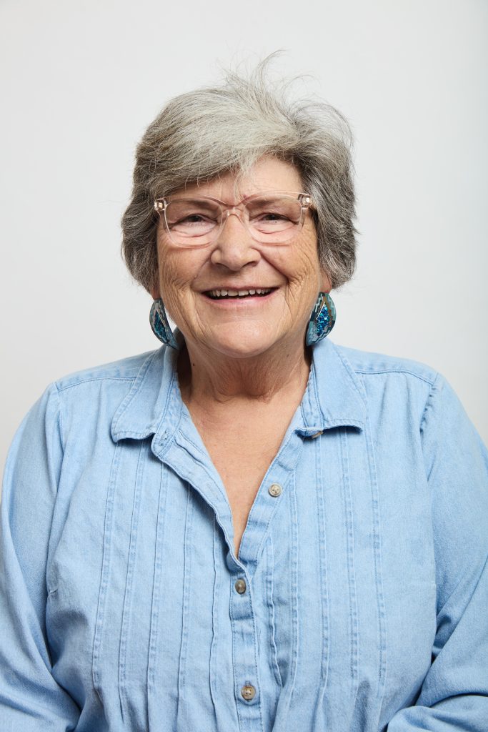 Smiling older woman with short gray hair and glasses wearing a light blue shirt and earrings.