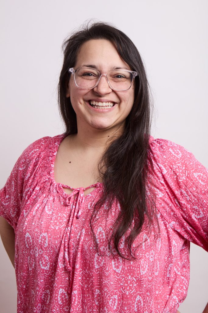 Smiling woman with long dark hair and glasses wearing a pink patterned top.