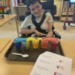 A student in their wheelchair sitting at a table with cups of different colored liquids.