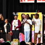 Five students stand together in matching t-shirts, singing on a stage. Three of them are wearing reindeer antlers. There are two teachers on stage. One teacher holds a mic for the student in the middle and the other holds the hand of a student.