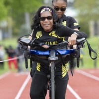 Kimar had tried several schools before being referred to MSB to find support for her Cortical Visual Impairment (CVI). At MSB, she quickly excelled after staff determined there needed to be a shift in her educational goals. From there, she became passionate about learning, and she found a love for sports. With MSB’s many mobility tools, Kimar has participated in cheerleading, track, and soccer. Best of all, she has found lifelong friends in her teammates and mobility skills that will help her enjoy even more exciting activities in the years to come. Two individuals on a track; the person in front is using an adaptive tricycle, assisted by the person behind. Both are wearing athletic gear and sunglasses, with others in the background.