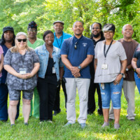 A group of eleven people stands outdoors on grass, smiling at the camera. Trees are visible in the background. A group of eleven people stands outdoors on grass, smiling at the camera. Trees are visible in the background.