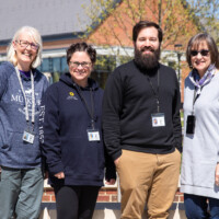 Four adults, two women and one man, wearing lanyards and standing outdoors, smiling in front of a building on a sunny day. Four adults, two women and one man, wearing lanyards and standing outdoors, smiling in front of a building on a sunny day.